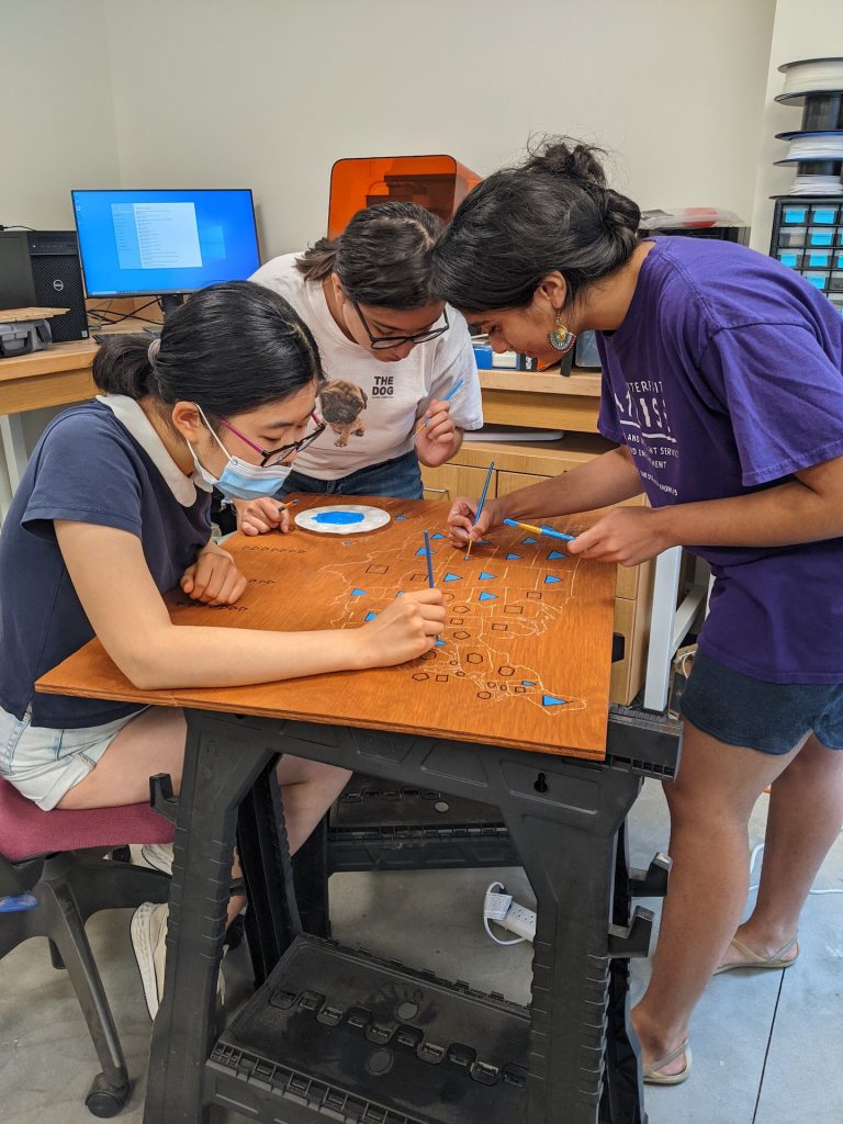 Three students add paint to a wooden map in the Makerspace